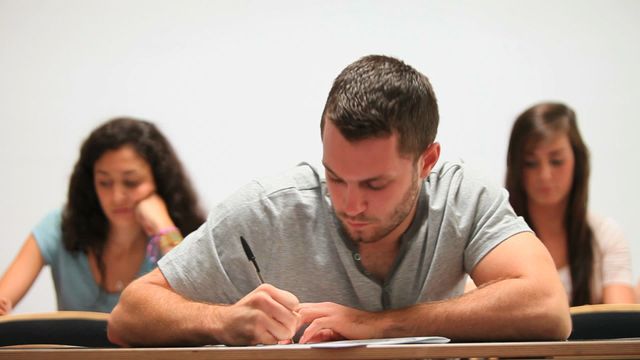 Young male student in a college classroom writing notes in a notebook while focusing intently. Background includes other students focusing on their tasks. A perfect visual for educational websites, academic publications, and study-related articles.