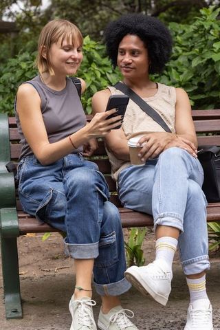 Couple sitting on park bench sharing smartphone and coffee, casual rolled-jeans lifestyle