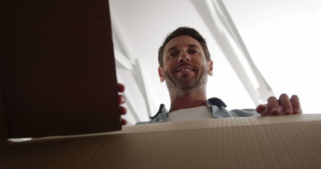 Man seen from below unpacking a cardboard box. Man appears to be in casual attired and is smiling. Ideal for concepts related to moving, relocation, delivery, and unpacking new items.