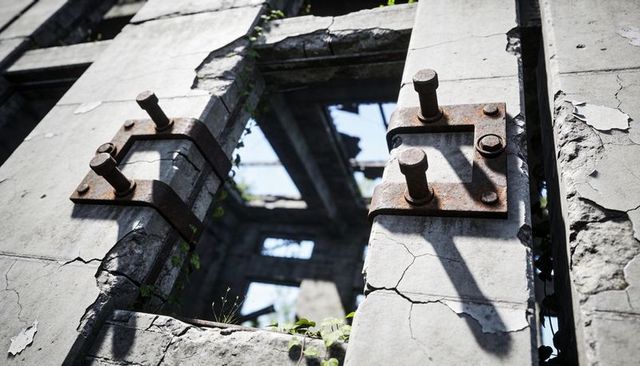 Rusting steel brackets clamping cracking concrete facade with vines and bolt detail