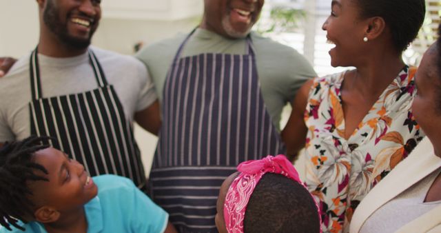This depicts an African American family enjoying their time together in the kitchen. Everyone is smiling and wearing aprons while having a joyful conversation, suggesting a strong bond and happiness. Perfect for advertisements or campaigns promoting family values, cooking together, home life, or community events.