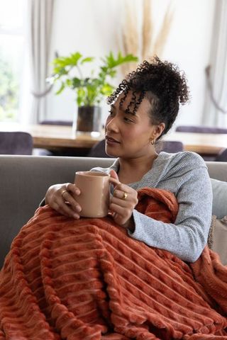 African american woman relaxing at home with rust-orange blanket and mug on cozy sofa