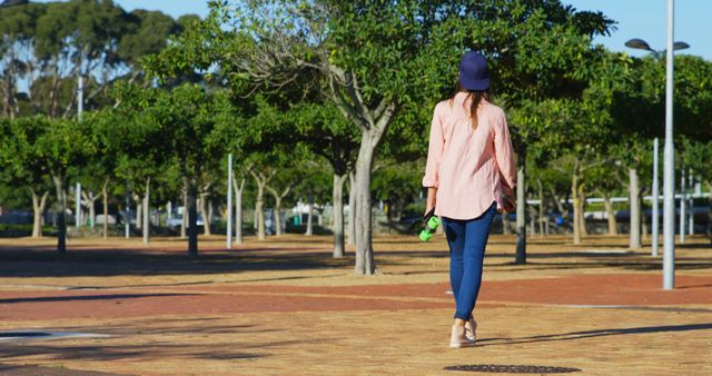 Woman walking on paved walkway in park holding skateboard. Wearing casual clothes and blue cap. Sunny weather with trees in background. Ideal for content related to outdoor activities, recreation, leisure, and lifestyle.