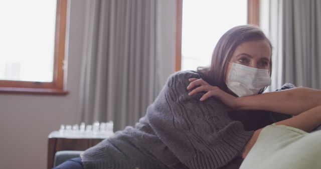 Woman wearing face mask leaning on couch at home during daytime - Download Free Stock Images Pikwizard.com