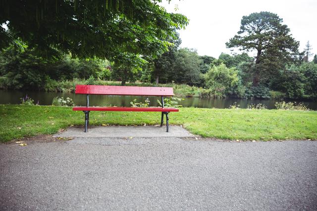 Empty red wooden bench by lakeside in park with lush greenery and trees in background. Ideal for themes of relaxation, solitude, nature, and peaceful outdoor settings. Suitable for use in travel brochures, nature blogs, and wellness websites.