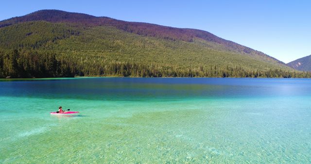 Solo kayaker paddling on peaceful, clear mountain lake surrounded by forested hills during summer. Perfect for promoting outdoor recreation, travel destinations, adventure vacations, or relaxation content.