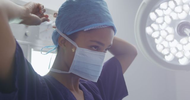 This image showcases a female surgeon in an operating room preparing for surgery by adjusting her cap and face mask. It can be used in medical publications, healthcare marketing materials, or educational resources aiming to highlight the professionalism and focus in medical environments. Suitable for articles on surgical procedures, gender equality in medicine, and hospital promotional content.