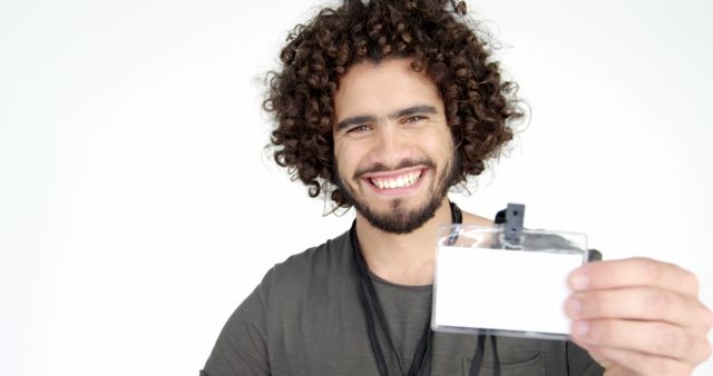 A young Caucasian man with curly hair is smiling and holding a name badge towards the camera, with copy space. His cheerful expression and the badge suggest a friendly introduction or a casual business environment.