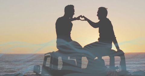 Mature couple making heart gesture sitting on suv roof at ocean sunset