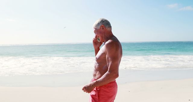 Senior man in red shorts is talking on his mobile phone while walking on the beach during a sunny day. The ocean waves can be seen in the background, conveying a relaxed and leisurely atmosphere. This image is ideal for use in advertisements focusing on senior lifestyle, beach vacations, and telecommunications services.