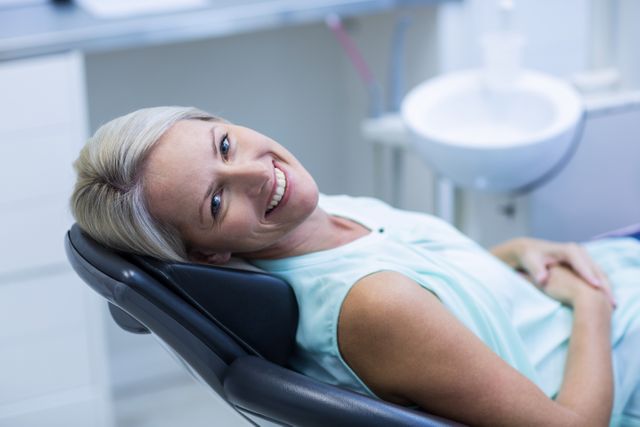 Blonde woman smiling while reclining in a dentist chair in a modern dental clinic. Ideal for use in healthcare promotions, dental care advertisements, patient comfort campaigns, and medical office brochures.