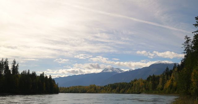 This image depicts a tranquil river flowing with mountains in the background and a sky dotted with clouds. Trees line the landscape, showcasing autumn colors. Perfect for use in nature-themed websites, travel brochures, or environmental campaigns highlighting serenity and the beauty of natural landscapes.