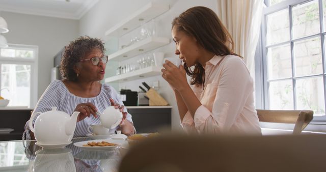 This image shows a grandmother and her adult granddaughter sharing a moment together while having tea at home. They appear to be conversing warmly around a table in a well-lit kitchen setting. This can be used to represent family bonds, intergenerational communication, homey moments, and familial love in advertisements, articles, or blogs about family life and relationships.
