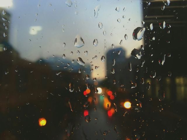 Rain drops on window create blurred background with colorful city lights at night. Captures peaceful evening mood in urban setting, great for website backgrounds, blogs about city life, or calming atmosphere designs.
