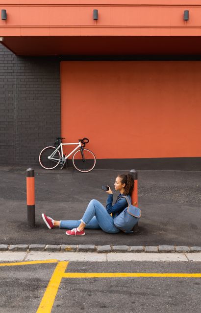 Full length of smiling young female biracial woman talking on mobile phone while sitting at sidewalk. unaltered, people, technology and city life concept.