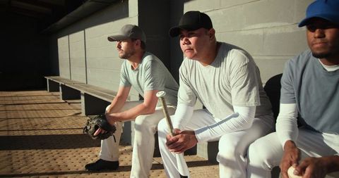 Baseball players sitting on dugout bench holding bat, glove and ball in sunlit dugout