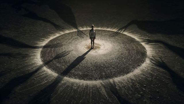 Solitary woman standing in glowing sand circle casting long shadows and reaching silhouettes