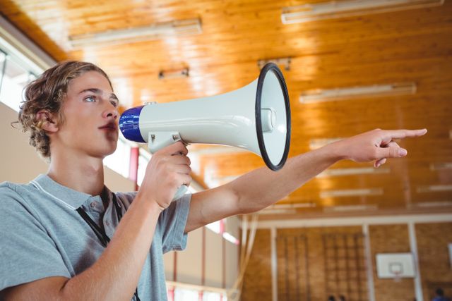 Male coach using megaphone in gymnasium, pointing and giving instructions. Ideal for concepts related to sports coaching, leadership, communication, and teamwork. Suitable for educational materials, sports training guides, and motivational content.