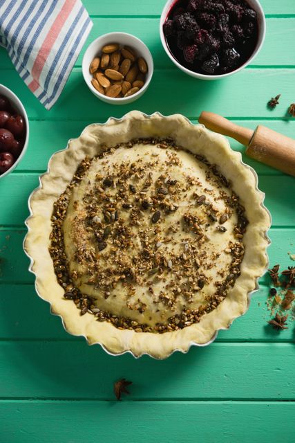 This image shows a homemade tart in the process of being prepared, with various spices and ingredients like almonds and berries on a green table. Ideal for use in cooking blogs, recipe websites, culinary magazines, and food-related advertisements.