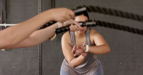 Women training with battle ropes checking smartwatch during partner workout in gym