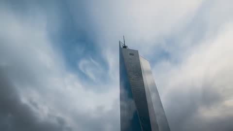 Glass skyscraper spire towering through drifting clouds with reflective curtainwall and moody sky
