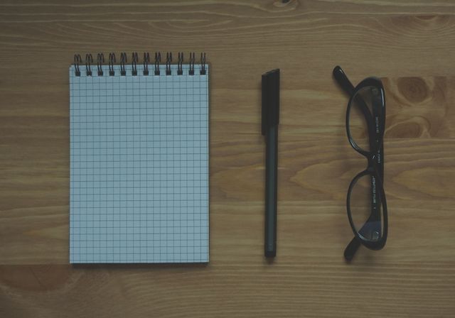 Flat lay of a minimalist workspace featuring a gridded notepad, black pen, and black-rimmed glasses on a wooden desk. Ideal for use in blog posts about productivity, educational materials, or office organization tips.