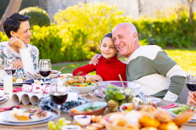 Family members of different generations are enjoying a meal together in a garden. The grandfather is hugging his granddaughter while the mother looks on with a smile. The table is filled with various dishes and drinks, creating a festive and joyful atmosphere. This image can be used for themes related to family bonding, outdoor dining, celebrations, and multigenerational relationships.