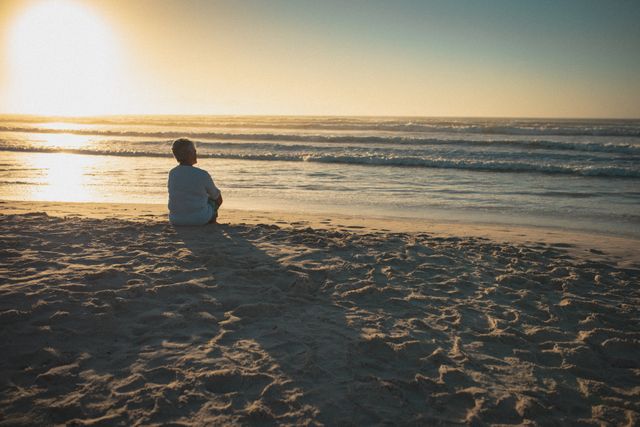 Senior woman sitting on sandy beach, gazing at ocean during sunset. Ideal for themes of relaxation, retirement, peaceful moments, and summer vacations. Can be used in travel brochures, wellness articles, and advertisements promoting beach destinations or retirement living.