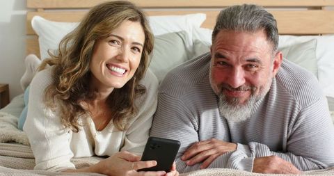 Middle-aged couple lying on bed smiling and holding smartphone, cozy bedroom lifestyle
