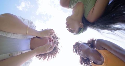 Three women leaning in circle framing bright sky, candid friendship outdoors
