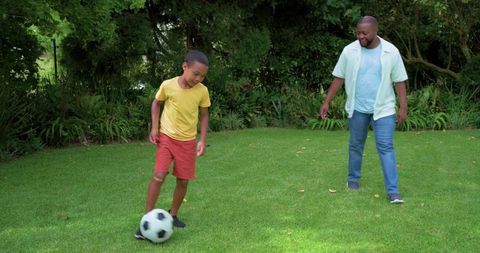 African american father coaching son playing soccer on green backyard lawn