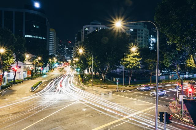 Long exposure of a busy city intersection captures light trails of vehicles passing through. Surrounded by tall buildings and illuminated by streetlights, this dynamic scene is perfect for urban lifestyle, travel advertisements, cityscape designs, and illustrating fast-paced city life.