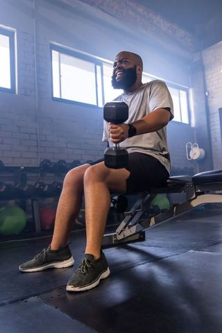 Bearded man lifting dumbbell while sitting on bench in industrial gym, intense workout