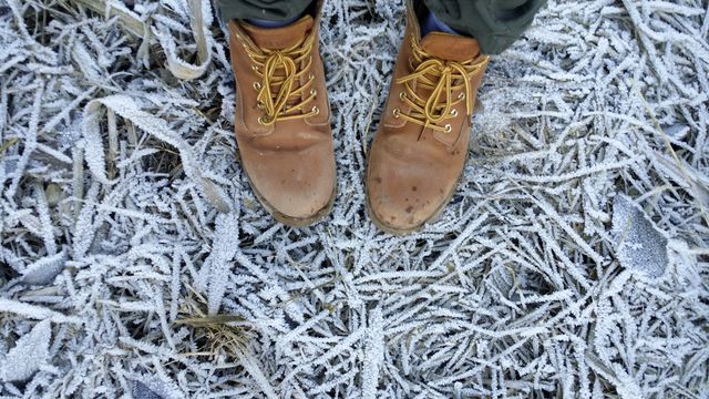 Close-up showing person wearing sturdy leather boots standing on grass covered with frost. Useful for depicting outdoor winter activities, durable footwear, winter landscapes, or exploring nature during cold weather.