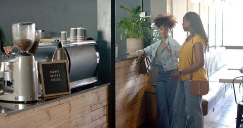 Two women chatting at coffee counter in sunlit cafe wearing denim and mustard top