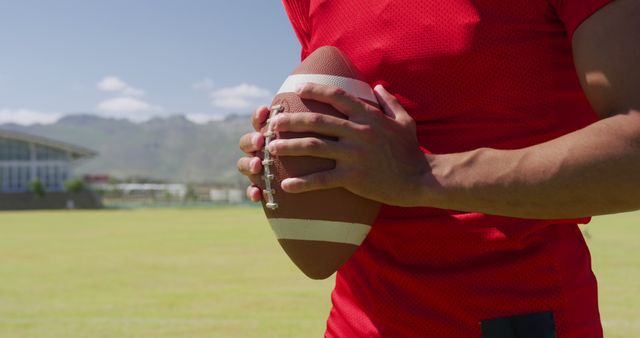Depicts a football player wearing a red jersey and holding a football on a sunny day in a large field. Ideal for sports-related content, articles about outdoor activities, athlete profiles, or promotional material for sports events.