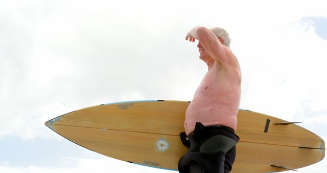 Senior surfer holding surfboard while looking at ocean, reflecting active lifestyle and passion for watersports. Suitable for use in advertisements or articles promoting active senior lifestyles, retirement activities, or health and fitness for older adults.