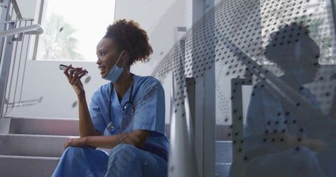 Nurse speaking into smartphone while resting on hospital stairs with mask and stethoscope