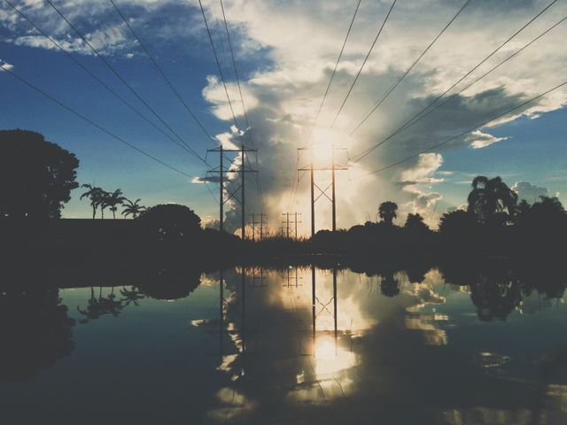 This image captures serene beauty combining natural and man-made elements. Tall power lines stretch across the sky as the sun sets behind clouds, casting a warm golden light. The reflection of the scene is perfectly mirrored on the calm water below, adding to the sense of tranquility. It is ideal for use in environmental awareness campaigns, energy and power industry materials, or as a calming nature-inspired decor.