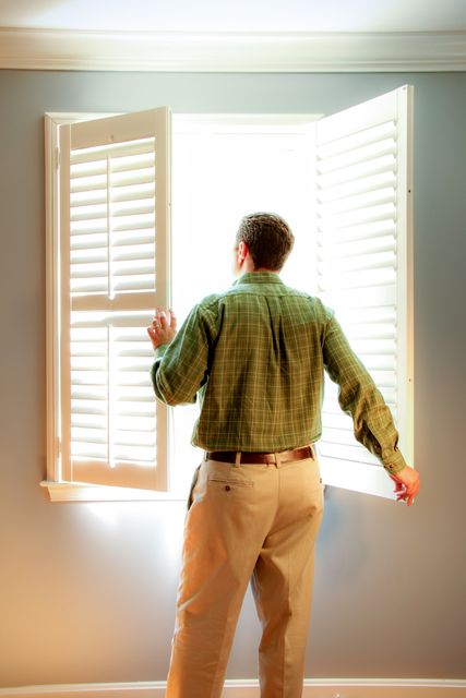 Man in green shirt standing by open window shutters, letting natural light into home. Ideal for themes of exploration, home living, fresh beginnings, and interior decor ideas.