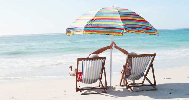 Elderly couple enjoying sunny day at the beach under a colorful umbrella, holding hands while sitting on lounge chairs. Perfect for illustrating vacations, retirement, leisure activities, travel brochures, summer holiday promotions, and lifestyle magazines.