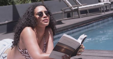Sunlit poolside reading: young woman lounging on deck with sunglasses and beachwear