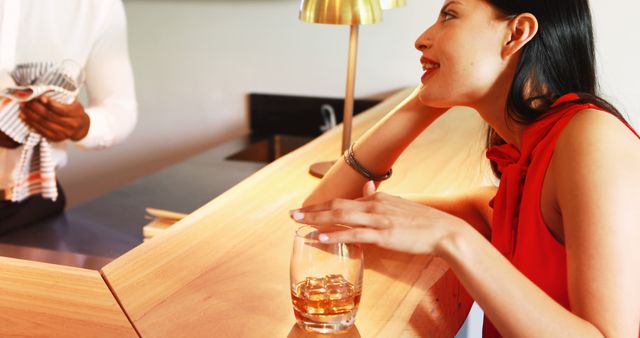 Woman in a red dress holding a glass of alcohol at a modern wooden bar counter. Suitable for images related to socializing, leisure activities, night out, bars, and lifestyle themes.