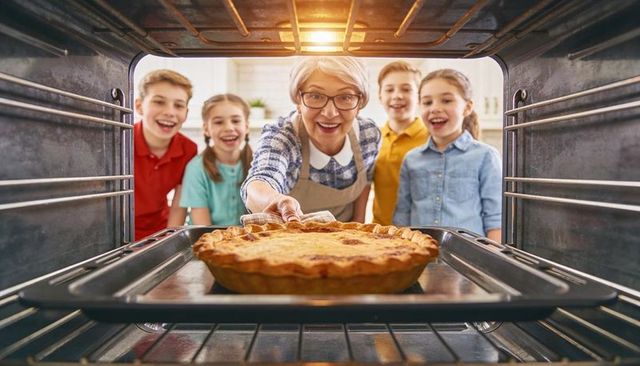 Senior woman pulling golden pie from oven while smiling grandchildren gather in cozy kitchen
