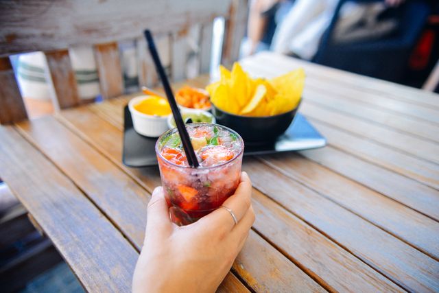 Hand holding a refreshing cocktail garnished with strawberries, with a bowl of nachos and assorted dips in the background on a wooden table. Perfect for use in promoting outdoor dining experiences, summer parties, social gatherings, and food and beverage establishments.