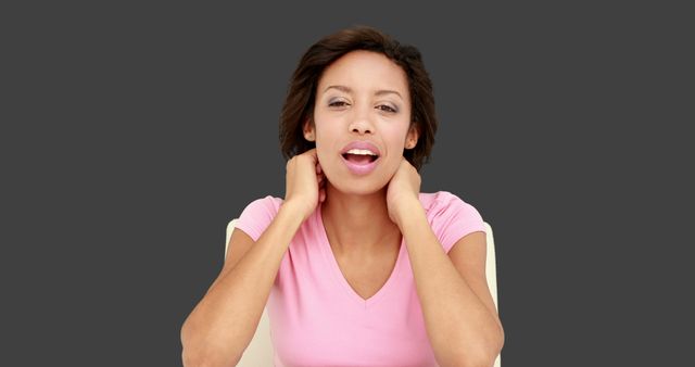 A mid-adult woman expressing tiredness by touching her neck. She is wearing a pink shirt and sitting on an isolated background. This image can be used in contexts related to exhaustion, fatigue, health problems, health and wellness blogs, symptom portrayal, and personal care tips.