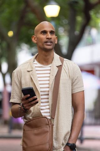 Indian man using smartphone on tree-lined urban plaza wearing brown crossbody bag