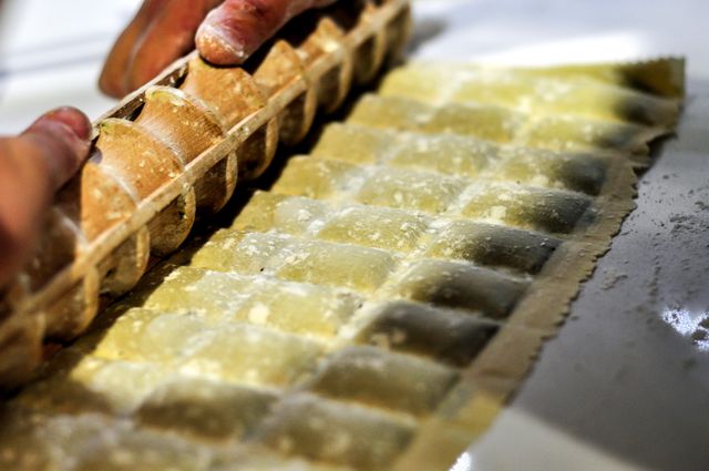 Hands are seen pressing ravioli dough with a specialized rolling tool, creating a pattern as the dough sheet lays on the counter. This image highlights the traditional process of homemade pasta-making, particularly ravioli. It is ideal for use in culinary blogs, recipe websites, or marketing materials for cooking classes focusing on Italian cuisine.