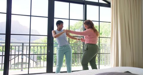 African american friends holding hands dancing beside sunlit balcony doors in bedroom