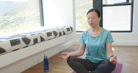 Asian woman meditating at home on yoga mat with earbuds and water bottle by window